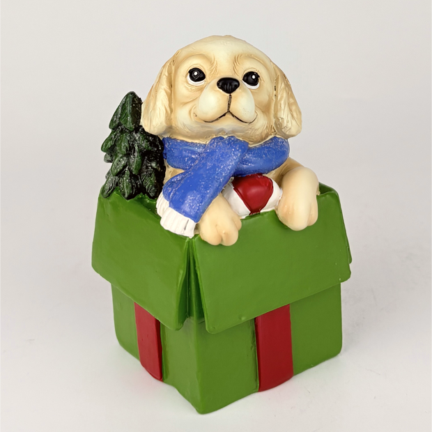 A decorative figurine of a cream-colored puppy wearing a blue scarf and holding a red heart, sitting inside a green gift box with red ribbon and a small black tree, on a light gray background.