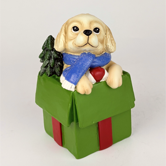 A decorative figurine of a cream-colored puppy wearing a blue scarf and holding a red heart, sitting inside a green gift box with red ribbon and a small black tree, on a light gray background.