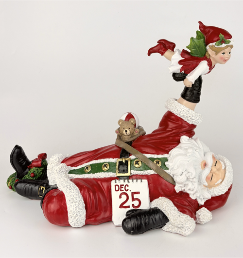 A Christmas figurine depicting Santa Claus lying down with a calendar showing Dec. 25, a teddy bear on his belt, and a small elf standing on his shoulder, all in red, white, and black with green holiday accents.