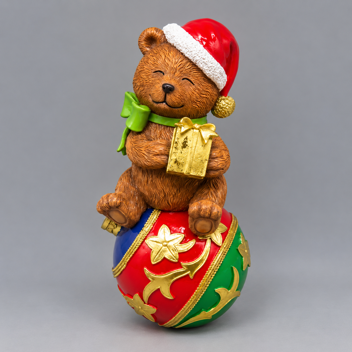 A festive Christmas-themed figurine featuring a brown teddy bear wearing a red Santa hat and green bow, sitting on top of a red, green, and gold ornament ball and holding a gold gift box, photographed against a neutral gray background.