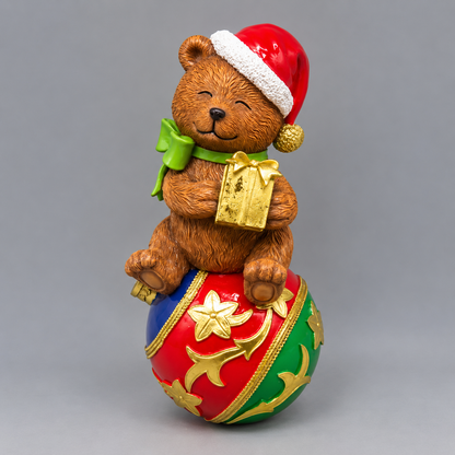 A festive Christmas-themed figurine featuring a brown teddy bear wearing a red Santa hat and green bow, sitting on top of a red, green, and gold ornament ball and holding a gold gift box, photographed against a neutral gray background.