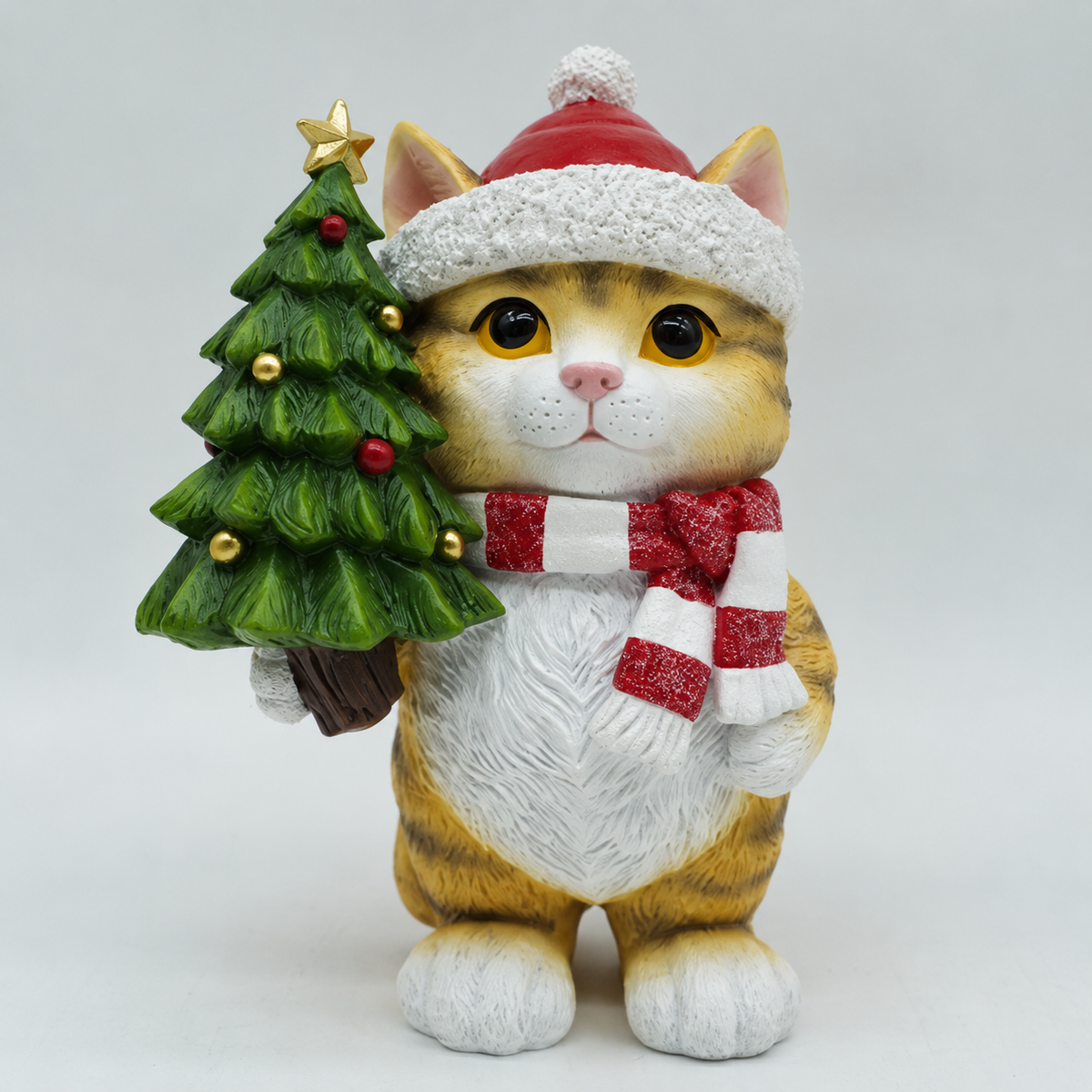 A festive cat figurine wearing a red and white Santa hat and matching striped scarf, holding a small decorated Christmas tree, photographed against a light gray studio background.