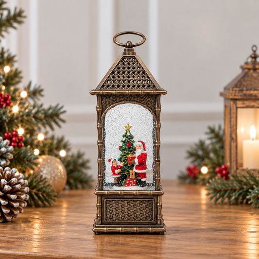 A decorative lantern-shaped Christmas ornament sits on a wooden table, featuring a miniature Christmas tree scene with Santa Claus inside. The lantern has an antique bronze finish and is framed by festive green pine branches with warm string lights in the background.