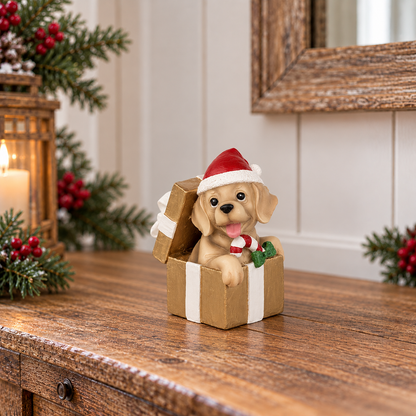 A decorative figurine of a golden retriever puppy sitting in an open gift box, wearing a red Santa hat and holding a candy cane, displayed on a wooden table with holiday decorations in the background.