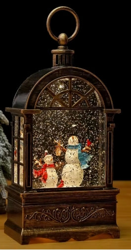 A decorative tabletop snow globe lantern with a dark brown carved wooden frame, a top hanging loop, and a clear dome showing two snowmen figures surrounded by falling white flakes. The product is photographed against a black background with a small blurred holiday tree prop at the left edge.