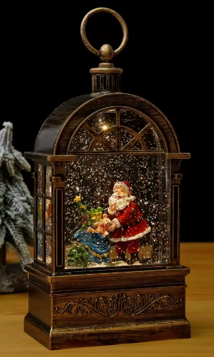 A decorative tabletop snow globe ornament shaped like a vintage arched window cabinet. It features a Santa Claus and child scene inside with falling snow effects, a hanging ring handle on top, and an ornate dark wood-look base, shown on a wooden surface against a black background.