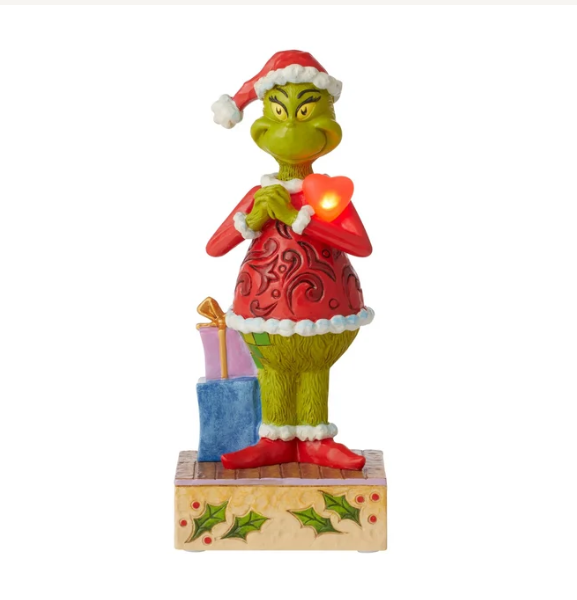 Grinch figurine dressed as Santa with a glowing red heart, standing on a decorated base with wrapped gifts against a white background.