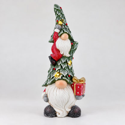 A festive Christmas gnome figurine with a tall, pointed green hat decorated with holly leaves, small yellow flowers, and a red Santa figure climbing it. The gnome has a long white beard, peach-colored hands, and holds a red gift box with a gold bow and polka dots.