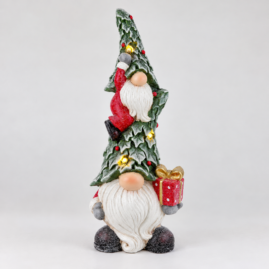 A festive Christmas gnome figurine with a tall, pointed green hat decorated with holly leaves, small yellow flowers, and a red Santa figure climbing it. The gnome has a long white beard, peach-colored hands, and holds a red gift box with a gold bow and polka dots.