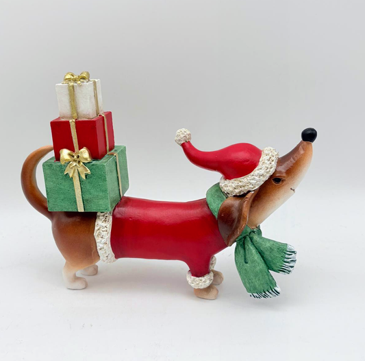 A dachshund figurine wearing a red Santa hat and green scarf, carrying a stack of three wrapped Christmas presents on its back, photographed against a plain light gray background.