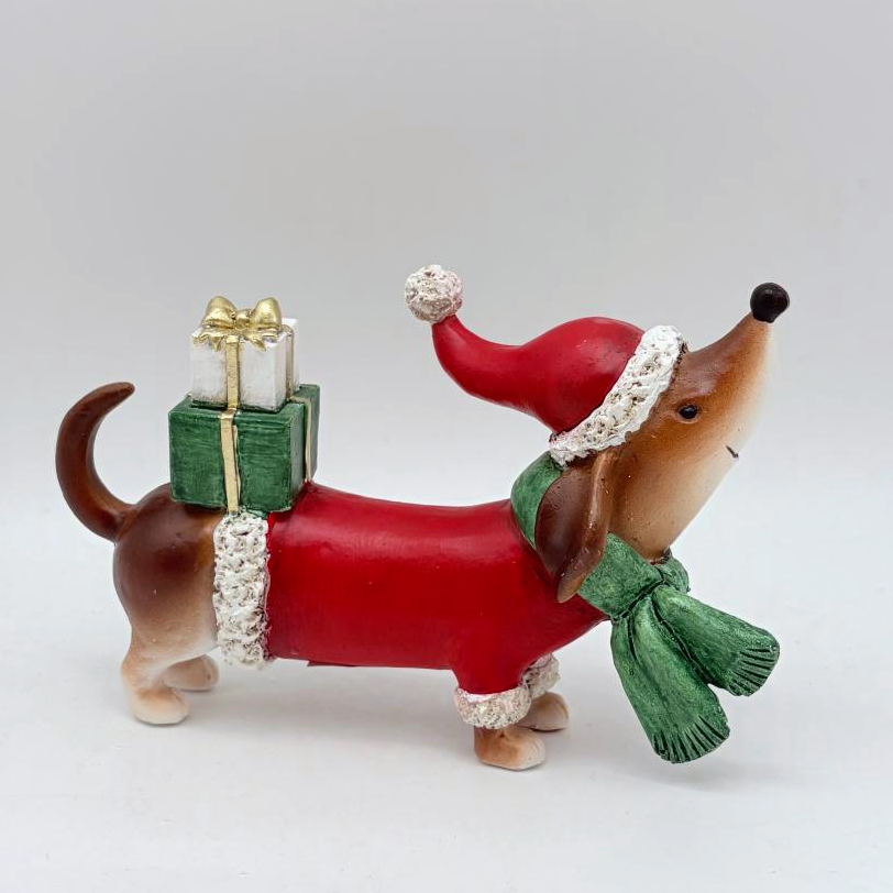 A dachshund figurine wearing a red Santa hat and green scarf, with a red body covering and a green gift box on its back, photographed against a plain light gray background.