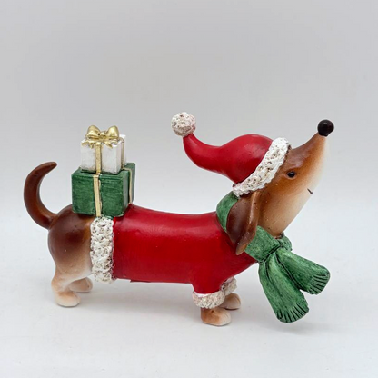 A dachshund figurine wearing a red Santa hat and green scarf, with a red body covering and a green gift box on its back, photographed against a plain light gray background.