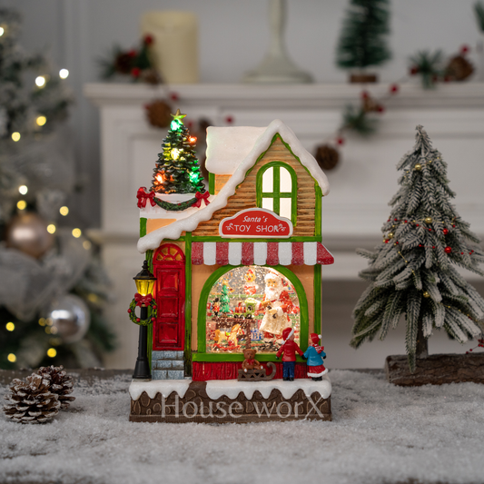 Decorative Christmas house snow globe with lights and Santa's Toy Shop sign, surrounded by festive decorations.