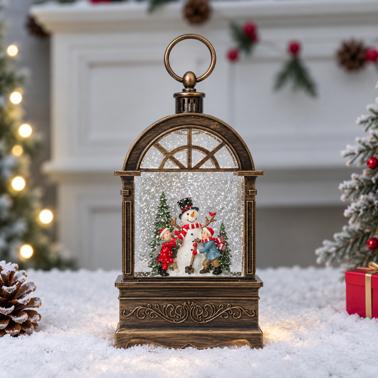 A decorative snow globe lantern with an ornate bronze-colored frame, featuring a snowman and Christmas tree scene inside, set on a snowy surface with a blurred holiday fireplace background.