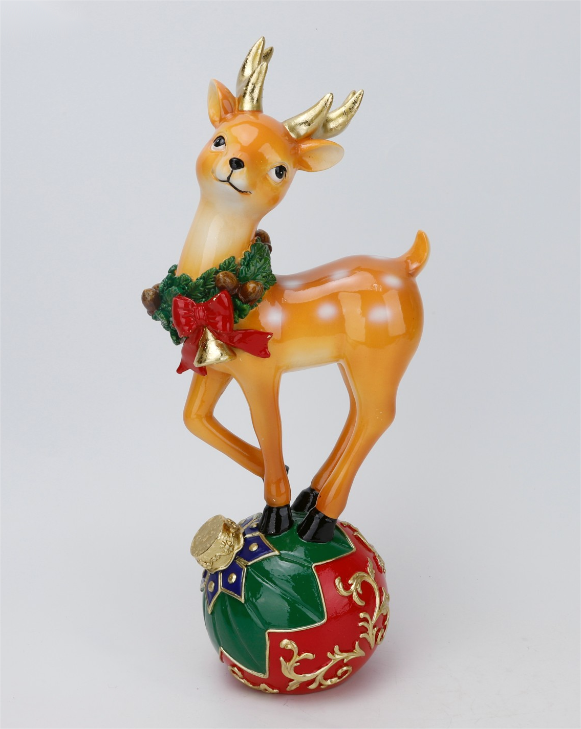 A festive Christmas ornament featuring a small golden deer wearing a green wreath with a red bow, standing on a red and green decorated ornament base against a light gray background.