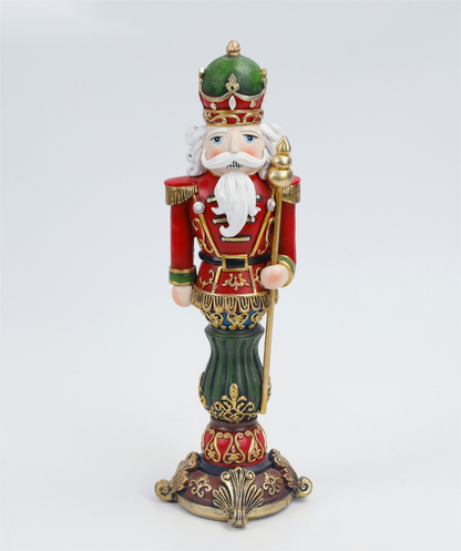 A detailed, hand-painted nutcracker figurine standing on an ornate base. The nutcracker is dressed in a red and green military uniform with gold trim, a white beard, and a green and gold crown, holding a gold staff.