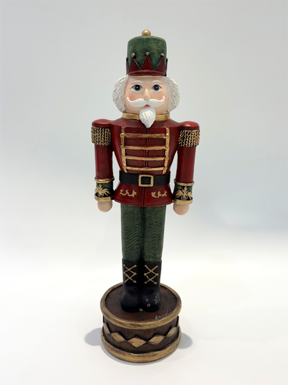 A traditional wooden nutcracker figurine standing on a round base, dressed in a red and green uniform with gold trim, white beard, and a green hat, photographed against a plain light gray background.