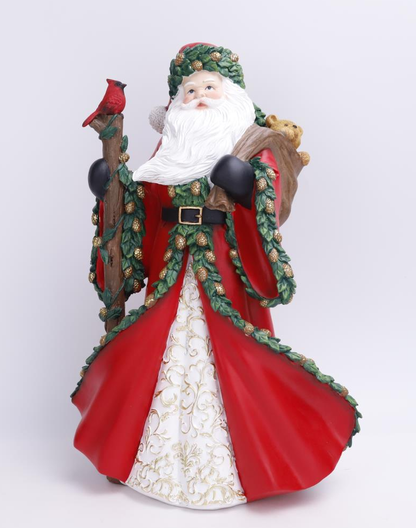 A Santa Claus figurine wearing a red coat with green pine garland trim, a white beard, and a brown sack on his shoulder, while holding a staff with a red cardinal perched on it, photographed against a light gray studio background.