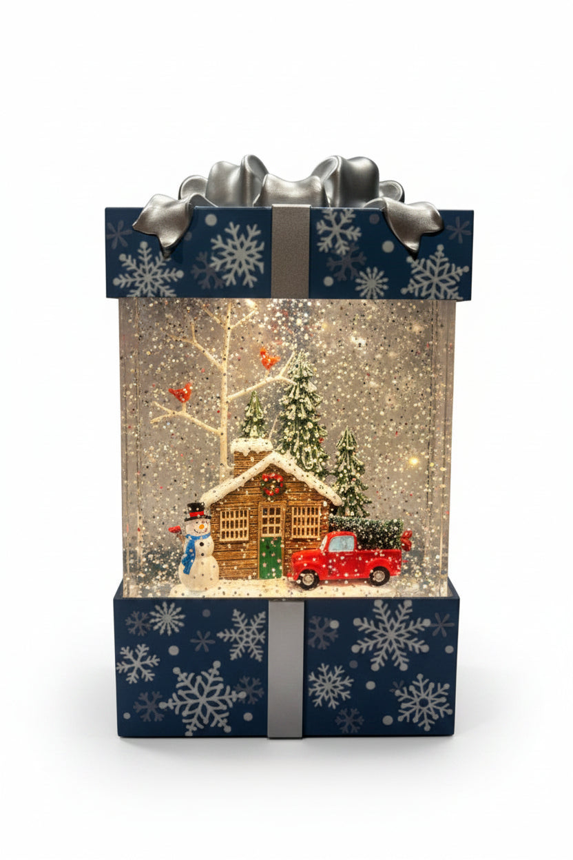 A decorative gift box snow globe Christmas light featuring a miniature winter scene with a glowing cabin, snowman, evergreen tree, red truck, and falling glitter, set inside a blue gift box with white snowflakes and silver bows.