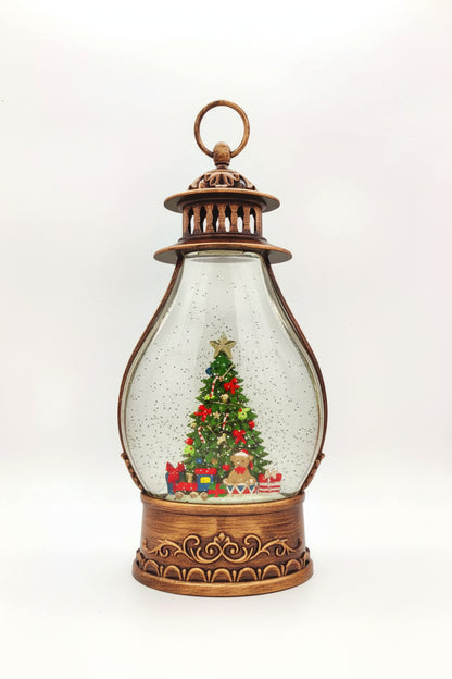 A vintage-style lantern Christmas snow globe decoration with an ornate bronze metal frame, clear glass body, and a miniature Christmas tree scene inside with gifts and snow.