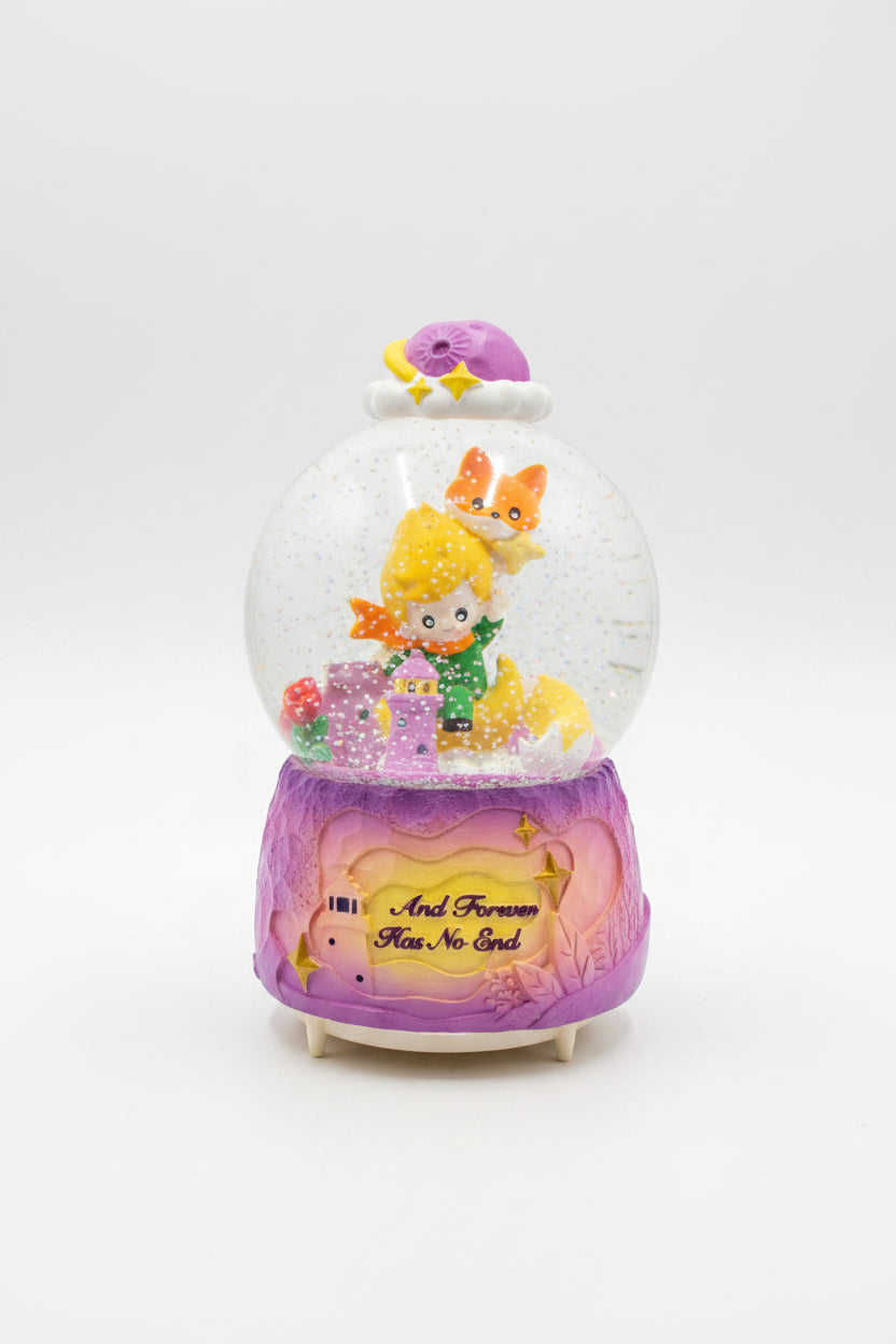 A pink and purple musical snow globe figurine featuring a cartoon girl and cat inside a clear dome, sitting on a dark granite countertop with a white box behind it.