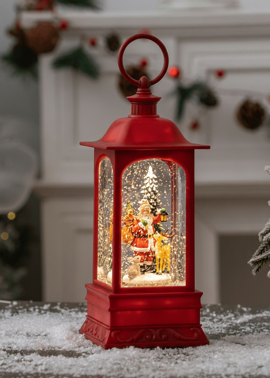 A red lantern-shaped Christmas snow globe sits on a snowy surface in front of a softly blurred holiday fireplace. The lantern has a hanging loop at the top and a miniature winter scene inside with Santa, a tree, and falling snow.