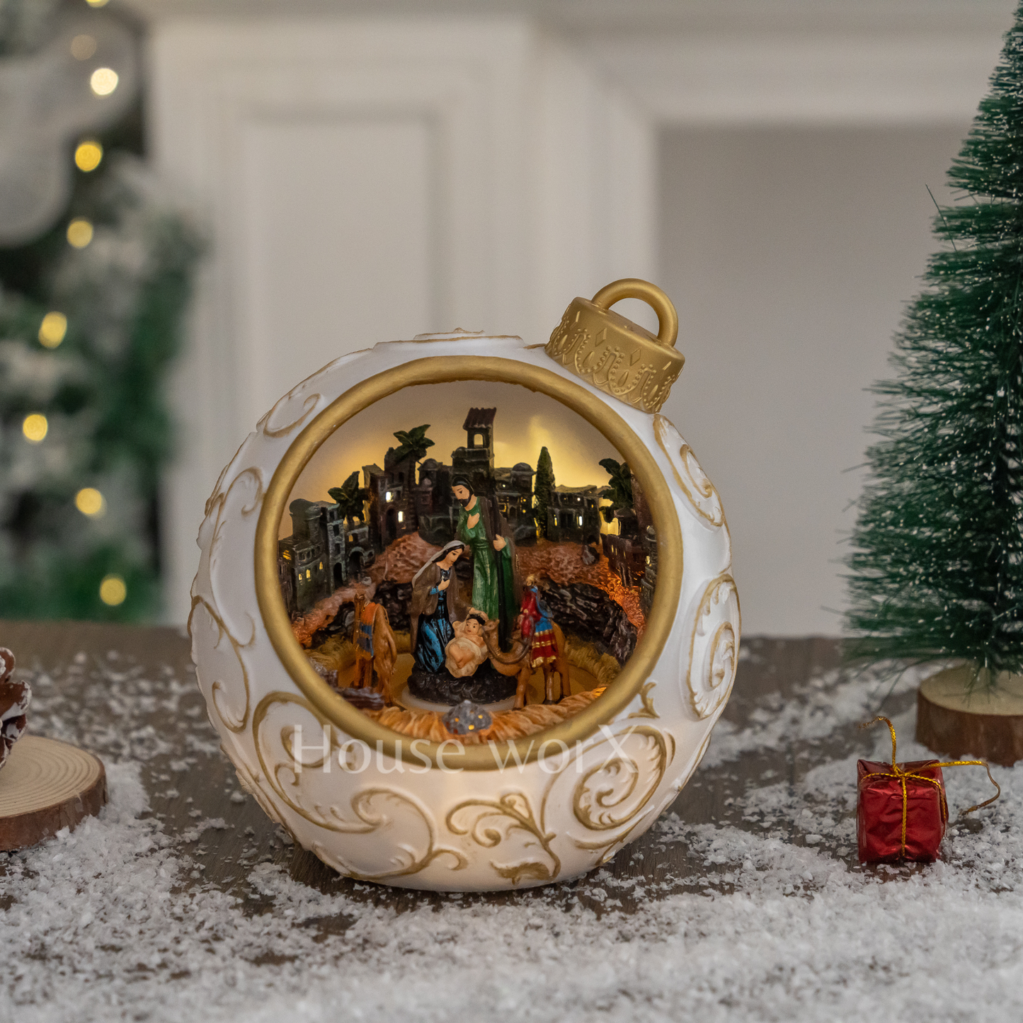 Decorative Christmas ornament with nativity scene inside, placed on a snowy surface with blurred Christmas trees in the background.