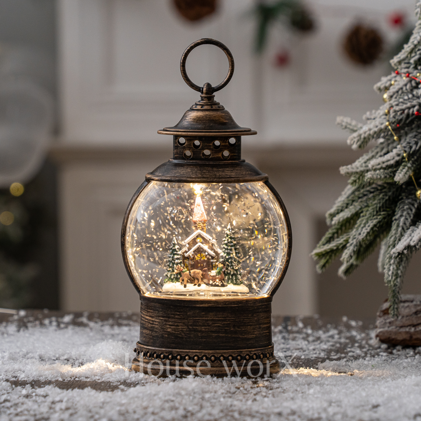 Decorative lantern with a snow globe inside, featuring a small village scene, on a surface with a blurred Christmas tree and decorations in the background.